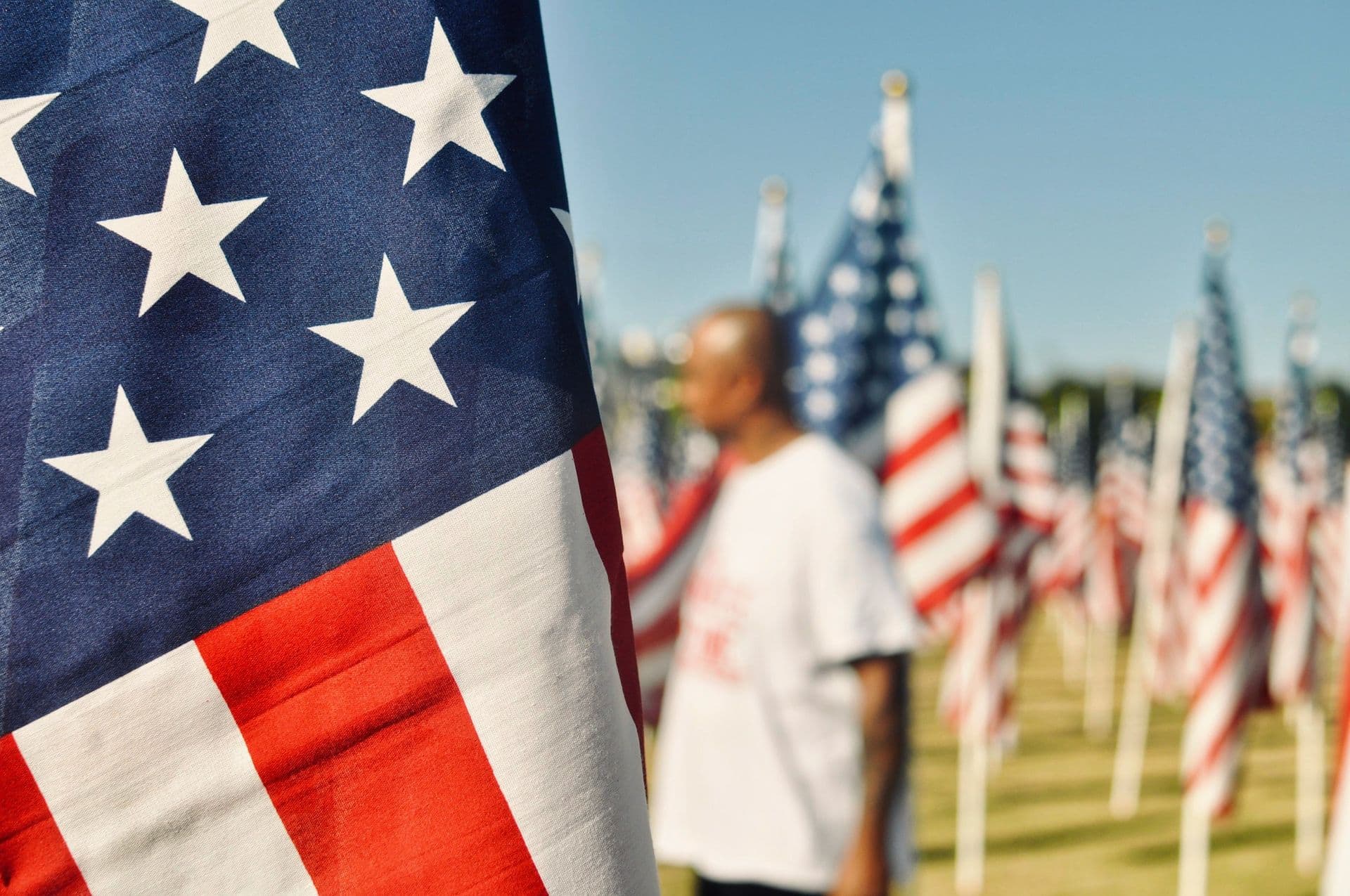 A closeup of an American flag in the foreground. In the background, a person stands among many more flags.
